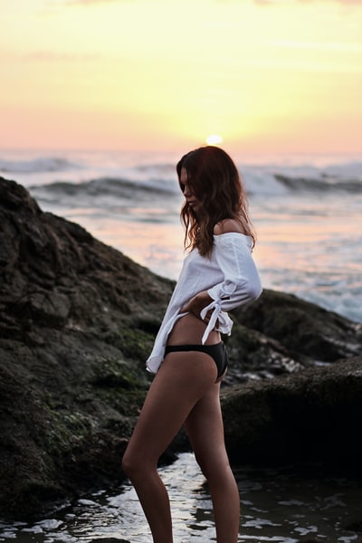 女人穿着白色了肩膀的衬衫和黑色的短裤站在岸上大岩石在金色的小时
